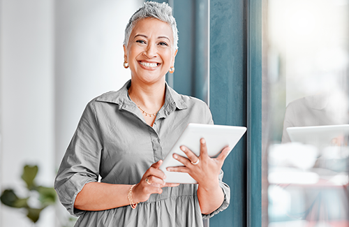 Professionally-dressed older woman smiles at the camera while holding a tablet and standing in a modern office space